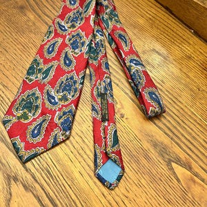May include: A red paisley silk tie with blue, green, and yellow accents. The tie features a repeating pattern of teardrop-shaped paisley designs. The tie is made of 100% silk.