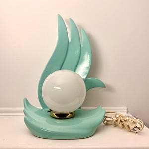 May include: A table lamp featuring a glossy, light turquoise ceramic base with curved, flame-like vertical accents. A spherical, opaque white glass globe sits in the center, secured by a gold-toned metal fixture with a white electrical cord.