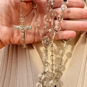 May include: A rosary featuring clear, faceted, round glass beads linked by silver-toned metal chain segments. The rosary includes a central oval medal depicting a religious figure and terminates in a silver-toned crucifix with a detailed figure.