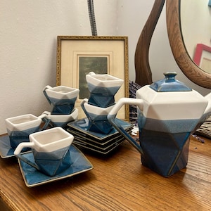 Puede incluir: Un juego de té geométrico con una tetera y varias tazas y platillos. El juego presenta un degradado de color blanco, azul claro y azul oscuro. Las tazas y los platillos son cuadrados, y la tetera tiene un diseño angular único.