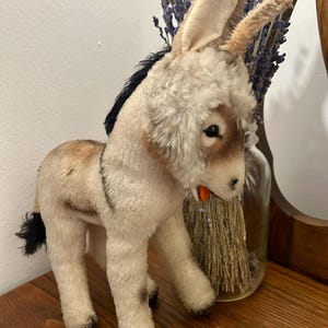 Op de afbeelding: Een vintage pluchen ezel speelgoed met een crèmekleurig lichaam, bruine accenten en een zwarte manen en staart. De ezel heeft een kleine oranje mond en staat op een houten oppervlak. Een klassiek kinderspeelgoed.