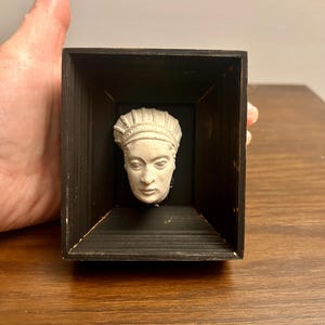May include: A small, off-white sculpted head bust, presented within a dark brown wooden box. The bust displays detailed facial features and a headdress. The rectangular box has a simple, minimalist design, with a dark brown finish.