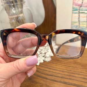 May include: Rectangular tortoiseshell-effect eyeglasses. The glasses are held by a hand with painted nails. The lenses are clear. The frame has a dark brown and amber colour.