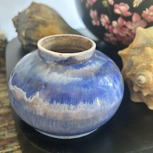 Peut inclure: Petit vase rond en céramique bleue à large ouverture. Le vase présente une finition texturée et émaillée avec des nuances de bleu et de blanc. Le vase est présenté avec des coquillages et un bol décoratif.