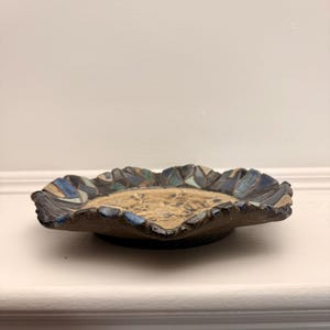 Könnte beinhalten: Eine flache Keramikschale mit einer strukturierten, beigen Mitte und einem dunklen, unebenen Rand, der mit einem mosaikartigen Muster aus blauen, türkisfarbenen und braunen glasierten Segmenten verziert ist. Der Rand ist gewellt und unregelmäßig.