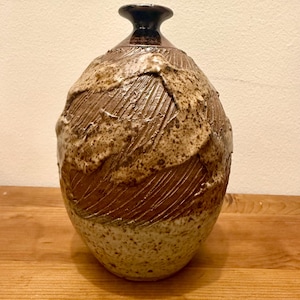 Puede incluir: Un jarrón de cerámica hecho a mano con un diseño texturizado y orgánico. El jarrón presenta un cuello y borde marrón oscuro, que transiciona a un cuerpo marrón claro y beige moteado. La superficie tiene líneas talladas y una estética áspera y terrosa.