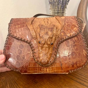 Könnte beinhalten: Eine braune Lederhandtasche mit einem Krokodil-Design auf der Frontklappe. Die Tasche hat einen Griff und eine strukturierte Oberfläche mit sichtbaren Nähten an den Rändern. Die Tasche ist aus braunem Leder gefertigt.