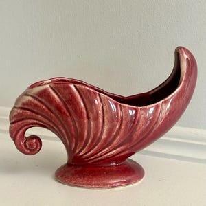 Puede incluir: Una maceta o recipiente decorativo de cerámica con un esmalte de color rojo intenso y crema, con una textura acanalada en forma de concha. El objeto tiene una silueta curva y alargada con un extremo elevado y una base circular.