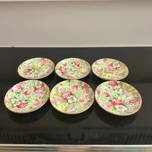 May include: Six small, oval plates with a floral design. The plates have a yellow base with pink, white, and green flowers. Each plate is edged with gold. The plates are arranged on a black surface, suitable for serving small portions.