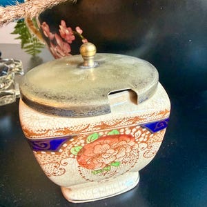 Puede incluir: Un azucarero de cerámica vintage con tapa de metal. El cuenco presenta un diseño floral en naranja, rojo y verde sobre un fondo crema, acentuado con ribetes azules y dorados. La tapa tiene un pequeño pomo.