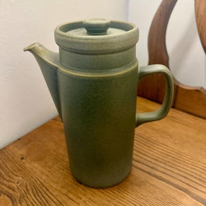 May include: A green ceramic coffee pot with a lid and handle. The pot has a cylindrical body and a spout for pouring. The color is a muted green with a matte finish. The pot is sitting on a wooden surface.