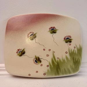 May include: A rectangular ceramic plate with rounded corners, featuring a cream-coloured base with a soft pink gradient at the top left. The surface is decorated with green painted grass, small red flower accents, and five stylised floral motifs.