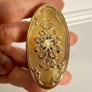 Könnte beinhalten: Ein ovales Kompaktgehäuse aus goldfarbenem Metall mit einem komplizierten, geprägten Schnörkelmuster auf der Oberfläche. Die Mitte des Gehäuses ist mit einem kreisförmigen Dekorelement verziert, das eine kleine weiße Kunstperle enthält.
