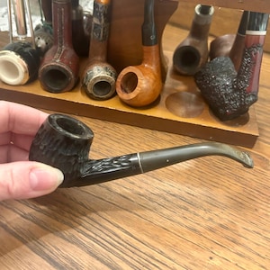 Peut inclure: Une pipe à tabac noire avec un foyer texturé et une tige incurvée. La pipe est tenue devant un présentoir à pipes en bois rempli de différentes pipes. Le présentoir est sur une surface en bois.