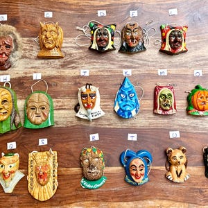 May include: A collection of twenty-four small, hand-painted decorative masks and figurines arranged in rows, each labeled with a letter. These items feature diverse faces, including mythical creatures, animals, and human-like characters in various colors.