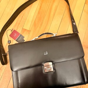 Puede incluir: Maletín de cuero negro con cerradura de combinación plateada y correa para el hombro desmontable. El maletín tiene un asa superior y un pequeño logotipo. Una etiqueta de marca está adjunta. El maletín mide 40 cm de ancho.