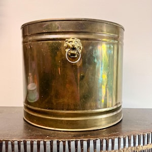 Puede incluir: Un cubo cilíndrico de latón con un acabado metálico pulido. El exterior presenta un adorno decorativo de cabeza de león centrado en la parte frontal, que sostiene un anillo metálico circular en su boca. El cubo tiene una superficie lisa y brillante.