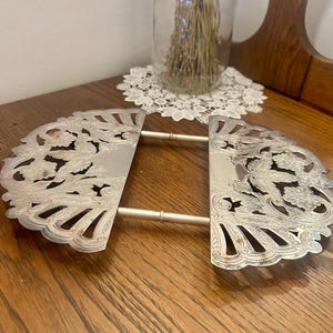 Peut inclure: Un dessous de plat extensible en métal argenté composé de deux plaques semi-circulaires aux motifs floraux découpés et aux bords festonnés. Les deux sections sont reliées par deux tiges métalliques coulissantes permettant le réglage.