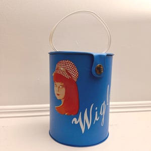 May include: A cylindrical blue wig storage case with a white handle and a snap closure. The front features a graphic illustration of a person with red hair wearing a red and white checkered hat, with the white cursive text "Wig" printed below.