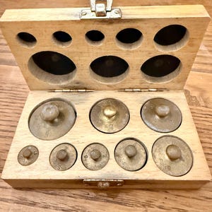 Op de afbeelding: Een vintage houten kist met een scharnierend deksel, met een set messing gewichten. De gewichten zijn van verschillende maten en vormen, met een knop aan de bovenkant. De kist heeft cirkelvormige uitsparingen om de gewichten vast te houden.