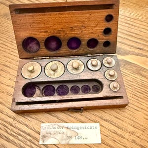 May include: A vintage wooden apothecary scale set with brass weights. The weights are labeled with numbers indicating their mass in grams. The set includes a wooden box with a hinged lid and a small paper label with text.
