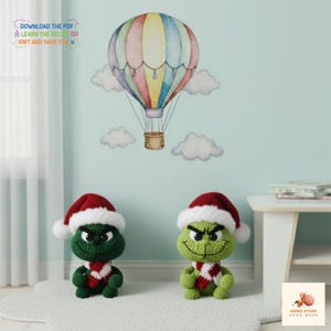 May include: Two green crochet figures wearing red and white Santa hats and scarves. A colorful hot air balloon and clouds are on the wall behind them. The text "DOWNLOAD THE PDF" and "KNIT AND HAVE FUN" is visible.