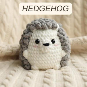 May include: A handmade crochet hedgehog in white and gray. The hedgehog has black eyes, a black nose, and pink cheeks. The word "HEDGEHOG" is written in a white rectangle above the hedgehog.