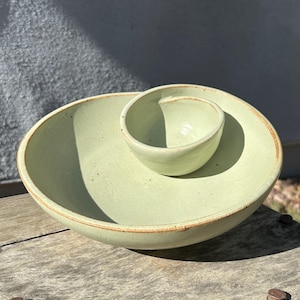 May include: A light green ceramic serving dish featuring a large, shallow circular bowl with an integrated smaller, deep bowl attached to the interior side. The piece has a smooth, glazed finish with subtle speckles and a raw, brown clay rim.