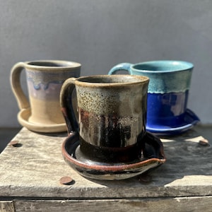 May include: Three ceramic mugs with integrated saucers. The front mug features a dark brown and black glaze with a speckled rim. Behind it, one mug has a light tan and lavender drip glaze, and the other has a vibrant blue and teal finish.