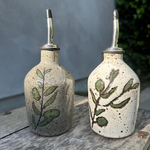 Peut inclure: Deux distributeurs d'huile en céramique avec becs verseurs en métal. La bouteille de gauche est gris clair avec des taches sombres et un motif de branche feuillue verte. La bouteille de droite est blanc cassé avec des taches brunes.