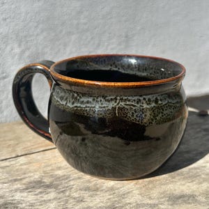 May include: A round ceramic mug with a glossy black finish and a wide handle. The upper portion of the mug features a textured, mottled band of white and dark green glaze, while the rim is accented with a thin, warm brown-toned edge.
