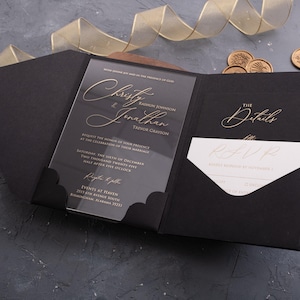 May include: Black wedding invitation with gold lettering on a clear acrylic card. The invitation includes details for the event, with a matching RSVP card in a black envelope. Gold ribbon and wax seals are also visible.
