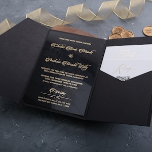 May include: Black wedding invitation with gold lettering. The invitation includes the names Chele Bené Woods and Andrew Harald Lilly, along with the date and time of the wedding. The invitation is in a black envelope with a pocket for an RSVP card.