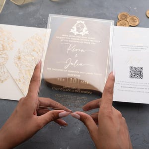 Könnte beinhalten: Transparente Hochzeitskarte aus Acryl mit goldenen Akzenten. Die Karte zeigt die Namen "Kevin & Julia" und das Datum "10. Februar 2024". Ein Umschlag und eine zusätzliche Karte sind ebenfalls zu sehen.