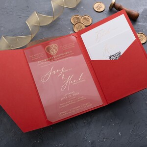 May include: A red wedding invitation suite with gold accents. The invitation features a clear acrylic overlay with gold calligraphy and a gold emblem. The suite includes a red pocket folder, inserts, and gold wax seals.