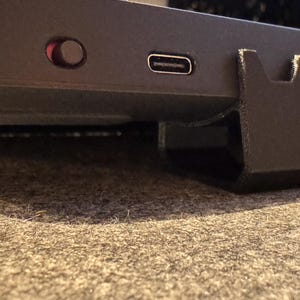 Puede incluir: Primer plano de un dispositivo electrónico negro con un puerto USB-C, un botón de encendido rojo y un soporte negro. El dispositivo está sobre una superficie alfombrada gris.