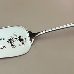 May include: Silver cake server with a long handle and a wide, oval blade. The blade features an engraved image of Mickey and Minnie Mouse kissing, with the names "Mike & Sanne" and the date "20-02-2026" below the image.