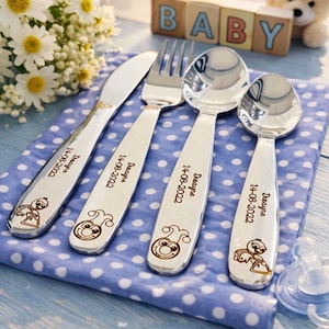 May include: A set of four personalized baby utensils: a knife, fork, and two spoons. Each utensil is engraved with the name "Dwayne" and the date "14-08-2022", along with a cartoon design. The set rests on a blue and white polka dot cloth.