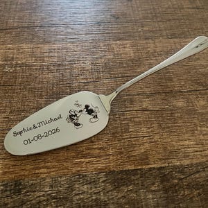 Op de afbeelding: Een zilverkleurige metalen taartschep met een gegraveerde illustratie van twee kussende tekenfilmmuizen, voorzien van de tekst "Sophie & Michael" en de datum "01-08-2026" op het platte, driehoekige blad van het keukengerei.