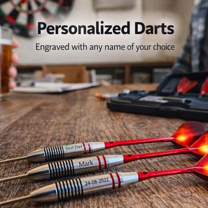 Op de afbeelding: Drie gepersonaliseerde darts met rode en zilveren accenten, gegraveerd met namen en data. De darts liggen op een houten tafel, met een vage achtergrond met een dartbord en een biertje. De tekst luidt "Gepersonaliseerde Darts".