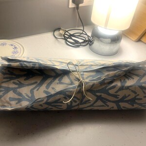 May include: A rectangular package wrapped in patterned paper with a blue coral design, tied with twine. The package is on a white countertop, with a plate and lamp in the background.