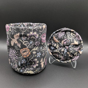 May include: A cylindrical fabric storage container and a matching circular fabric lid, both featuring a dark gray background with a repeating pattern of orange, purple, and light blue flowers, leaves, and small winged dragon figures.