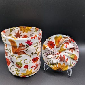 May include: A fabric storage basket and a matching circular fabric lid featuring a white background with a repeating pattern of orange squirrels, yellow sunflowers, red flowers, brown autumn leaves, green acorns, and small red mushrooms.