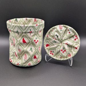 May include: A cylindrical fabric container and its matching circular lid, both featuring a cream-colored background with a green diamond lattice pattern. The design includes small red cardinal birds, green pine sprigs, and clusters of red berries.