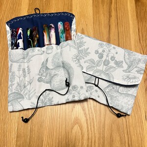 Peut inclure: Un étui à stylos en tissu avec une doublure bleue marine et un imprimé forestier gris clair avec des écureuils, des champignons et des fleurs. Le rouleau contient plusieurs stylos colorés. Des cordons noirs sécurisent le rouleau.