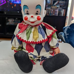 May include: A vintage clown doll with a bald head, painted facial features, and a colorful harlequin costume. The doll has large black shoes and is sitting on a white surface. The clown's face features red cheeks, a red nose, and a red smile.
