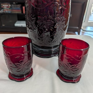 May include: A ruby red glass drink set featuring a large vase and two smaller glasses. The set has a floral design etched into the glass. The vase is approximately 8 inches tall, and the glasses are about 4 inches tall.