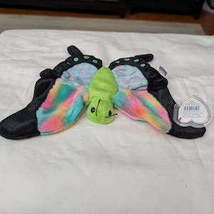 May include: A plush butterfly toy with black wings and colorful rainbow-colored accents. The butterfly's body is green, and it has a tag attached. The toy is lying on a white surface.