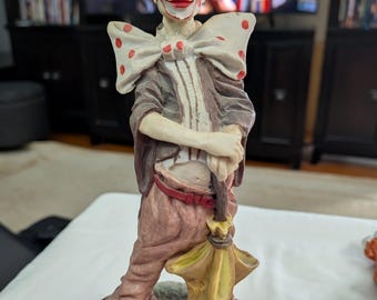 May include: A ceramic clown figurine with a green hat, red nose, and a large polka-dot bow tie. The clown wears a brown jacket, pink pants, and holds a yellow umbrella. The figurine stands on a wooden base.