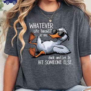 May include: Dark gray t-shirt featuring a cartoon duck wearing sunglasses, with the text "WHATEVER Life THROWS AT YOU, duck and Let It HIT SOMEONE ELSE." The duck is white with orange feet and beak.